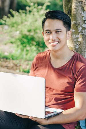 young male student with laptop smiling while studying online under the tree in the parkの写真素材