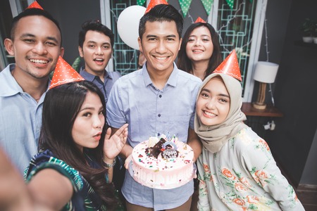 portrait of happy young asian people celebrating birthday of their friends together at home and taking selfieの写真素材