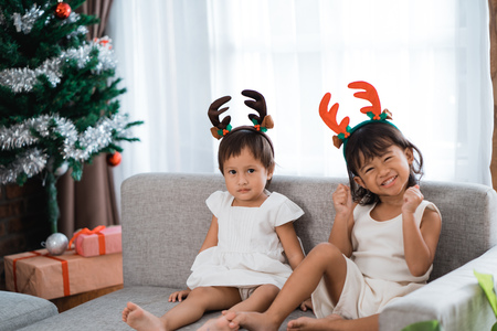 portrait of togetherness two daughter sitting on a couch wearing christmas headbandの写真素材