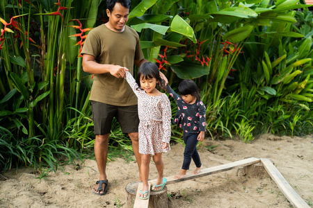 portrait a father helping two daughter with holding his hands when playing balance beamの写真素材