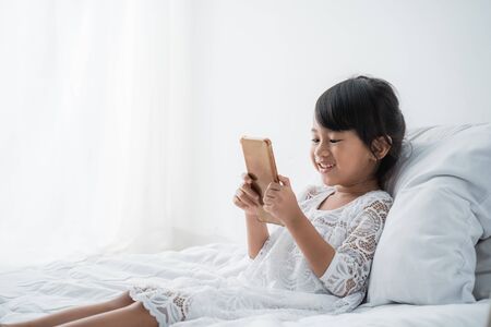 kid lying on bed wearing white using mobile phoneの写真素材