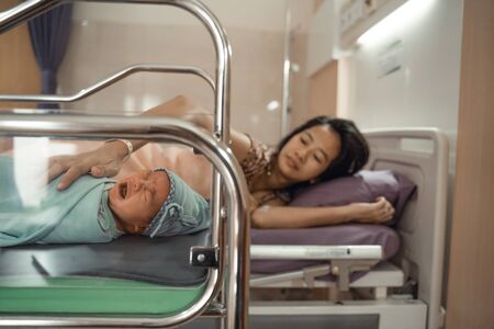 Crying newborn baby laying in crib with his mother lying on bed sideの写真素材