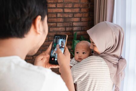 father taking photo of his baby infant at home while being carried by motherの写真素材