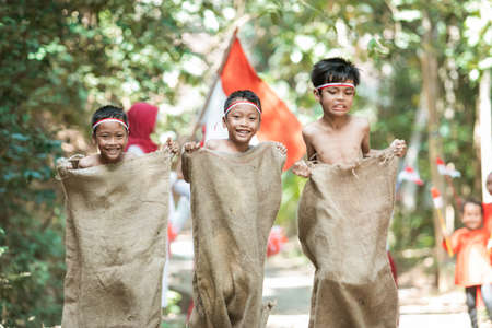 three children try to run fast and jump in the sack race with friends support themの写真素材
