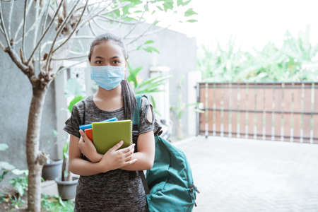a teenage girl standing wearing a mask and backpack carrying a book outdoorsの写真素材