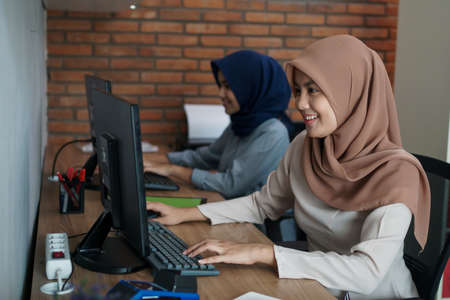 attractive cheerful young muslim business woman working on computer and smiling while sitting at her desk modern office with her friendの写真素材