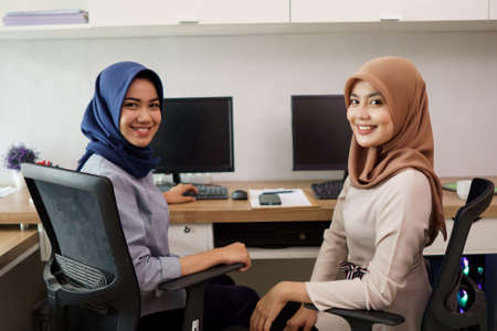 attractive cheerful young muslim business woman working and smiling while sitting at her desk modern office with her friendの写真素材