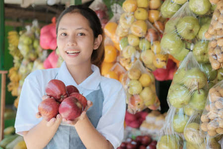 smiling asian lady shop assistant carrying fresh red apples on the background of a fresh fruitの写真素材