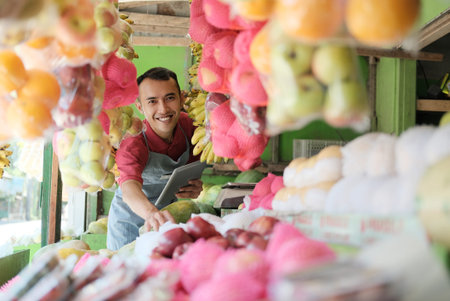 smiling Asian male stall waiter holding a digital tablet looking at the camera among the fresh fruit displaysの写真素材