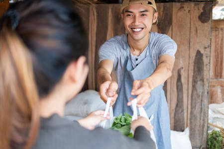 Close up of smiling Asian man holding plastic bags filled with vegetables as they are handed out to consumerの写真素材