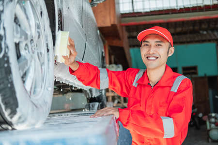 Male car cleaner wears red uniform and smiling hat while washing the bottom of the carの写真素材