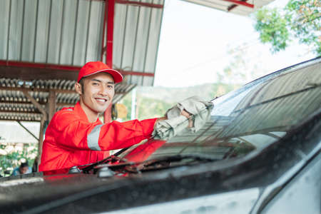 Asian male car cleaner wearing red uniform smiles while wiping car glassの写真素材