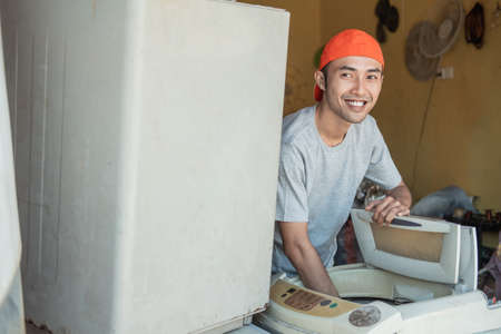 smiling asian male electronics worker looking at the camera as he unscrew the tube to fix a broken washing machine tubeの写真素材