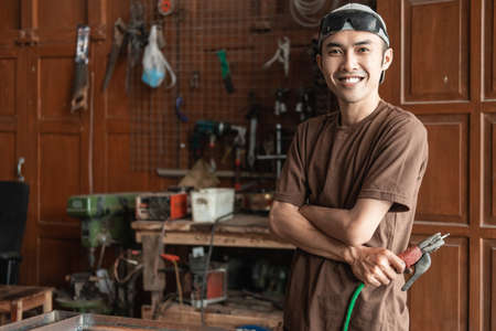 Male welder smiles with crossed hands while holding electric welderの写真素材