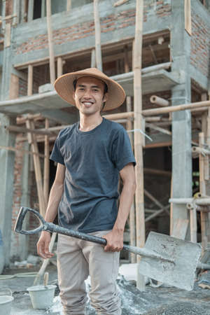 An Asian mason wearing a smiling hat stands up with a shovelの写真素材
