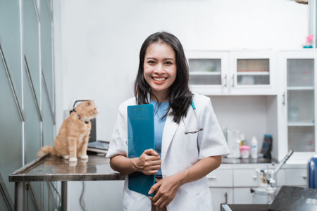 asian veterinarian smiling at camera while holding a clipboard near a cat sits on the tableの写真素材