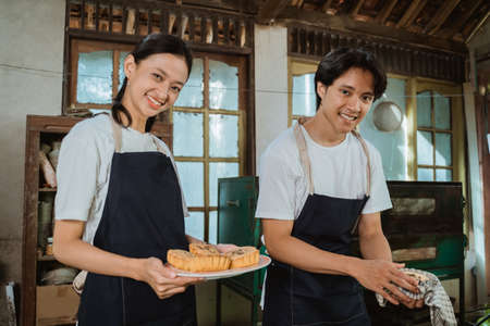 smiling young couple of cake makers wearing aprons holding up a tray of cooked cakesの写真素材