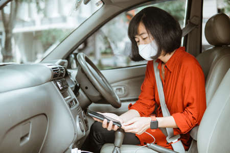 Woman in protective mask driving a car on road. charge phoneの写真素材