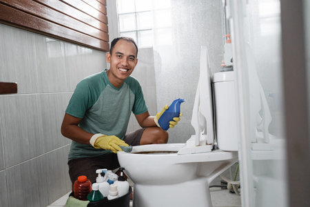 smiling male looks to camera hold sponge and a cleaning fluid bottle when cleaning the dirty toilet