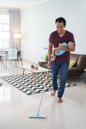 father cleaning the floor using broom while carrying his infant baby