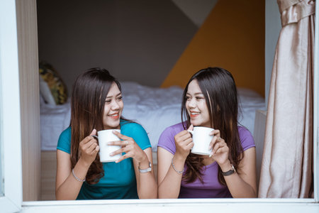 two twin Attractive asian sister holding a cup with hot tea or coffee near the windowの写真素材