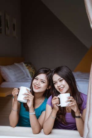 two twin Attractive asian sister holding a cup with hot tea or coffee near the windowの写真素材