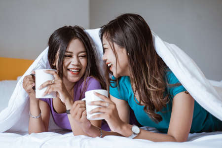 beautiful asian woman holding a cup with hot tea or coffee while laying on the bed togetherの写真素材