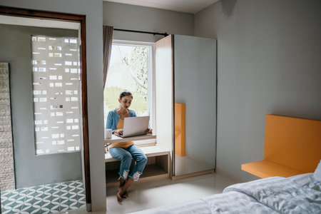 young woman in smart casual wear working on laptop while sitting near window at home