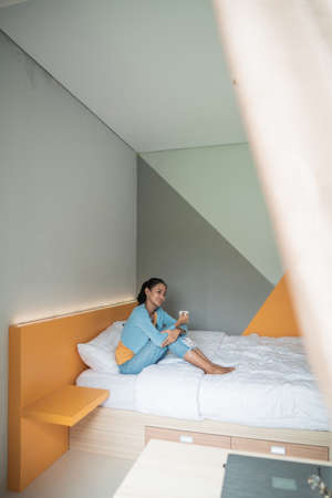 Attractive asian girl holding a cup with hot tea or coffee and looking at the window in her home and having a perfect cozy morning.の写真素材