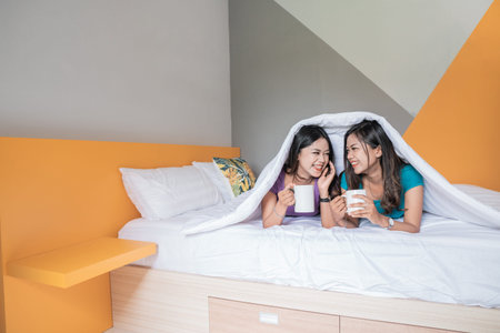 beautiful asian woman holding a cup with hot tea or coffee while laying on the bed together