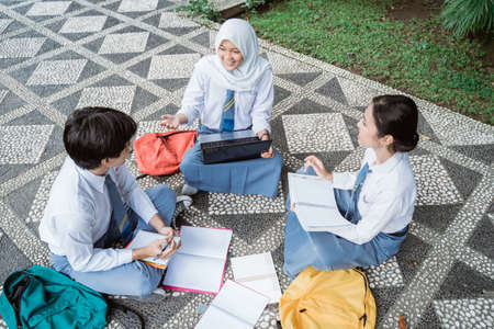 three high school students holding books and using laptops during group study with friendsの写真素材