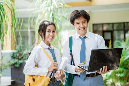 close up of a couple of high school students smiling at the camera while standing holding a book and wearing a bag while using a laptop computerの写真素材