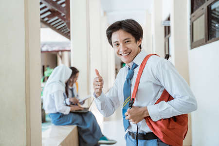 a high school boy smiles at the camera with thumbs up wearing a school bagの写真素材