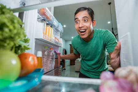 excited starving young asian man opening the fridge door looking for food to eatの写真素材