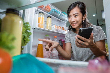 asian young woman using her smartphone to buy groceries while open her fridge at homeの写真素材