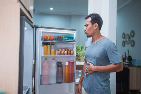 portrait of starving young asian man hold his belly while open the fridge door looking for something to eatの写真素材