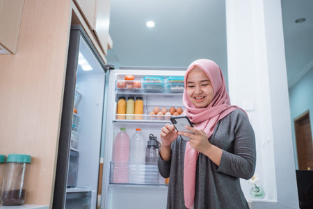 asian muslim young woman using her smartphone to buy groceries while open her fridge at homeの写真素材
