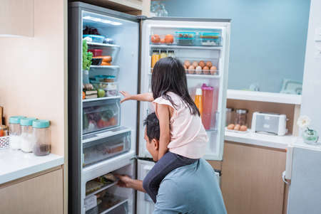 Young asian father and daughter open refrigerator at home looking for some foodの写真素材