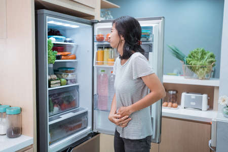 portrait of starving young asian woman hold her belly while open the fridge door looking for something to eatの写真素材