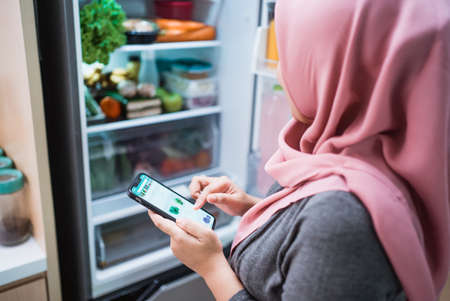 asian muslim young woman using her smartphone to buy groceries while open her fridge at homeの写真素材