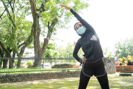 A pretty muslim woman athlete stretching and exercising her body outdoor and wearing face mask for...