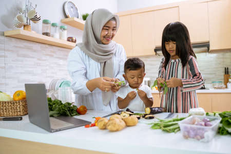muslim mother watching cooking video on laptop and making dinner with her two children in the kitchen togetherの写真素材