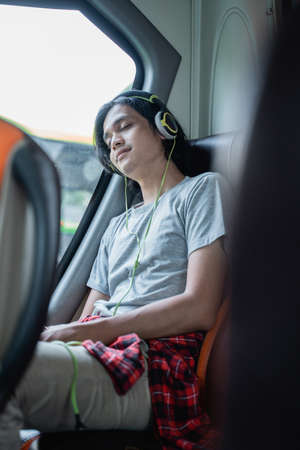 a young man wearing sleep headphones while listening to music while sitting by the window on a bus tripの写真素材