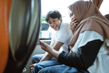 Asian Muslim couples laugh when they see the video on their cellphones while sitting on the busの写真素材