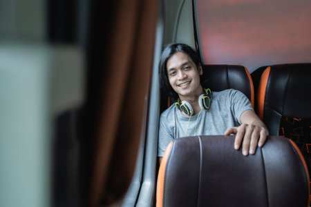 a young man in headphones smiles at the camera while sitting by a window on the bus
