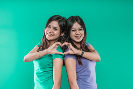 Beautiful twin sisters show a heart from their hands on a green background.の写真素材