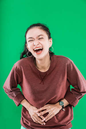 young Asian woman wearing sweater happy laughing while holding her stomach on green backgroundの写真素材