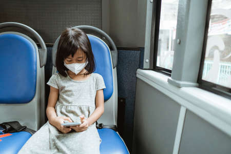 a little girl wearing a mask sits on a bench while using a smartphone on the bus while travelingの写真素材