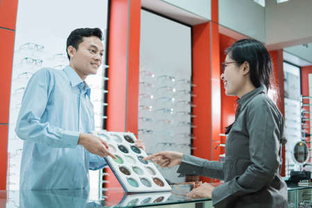 a male shop clerk holds a sample of eyeglass lenses and a customer points his finger to select eyeglass lenses at eyewear shopの写真素材