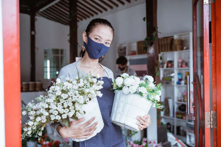 woman manager wearing face mask florist standing holding bucket flower with her staff in background. Fresh flowers in her floral shop following protocol healthyの写真素材